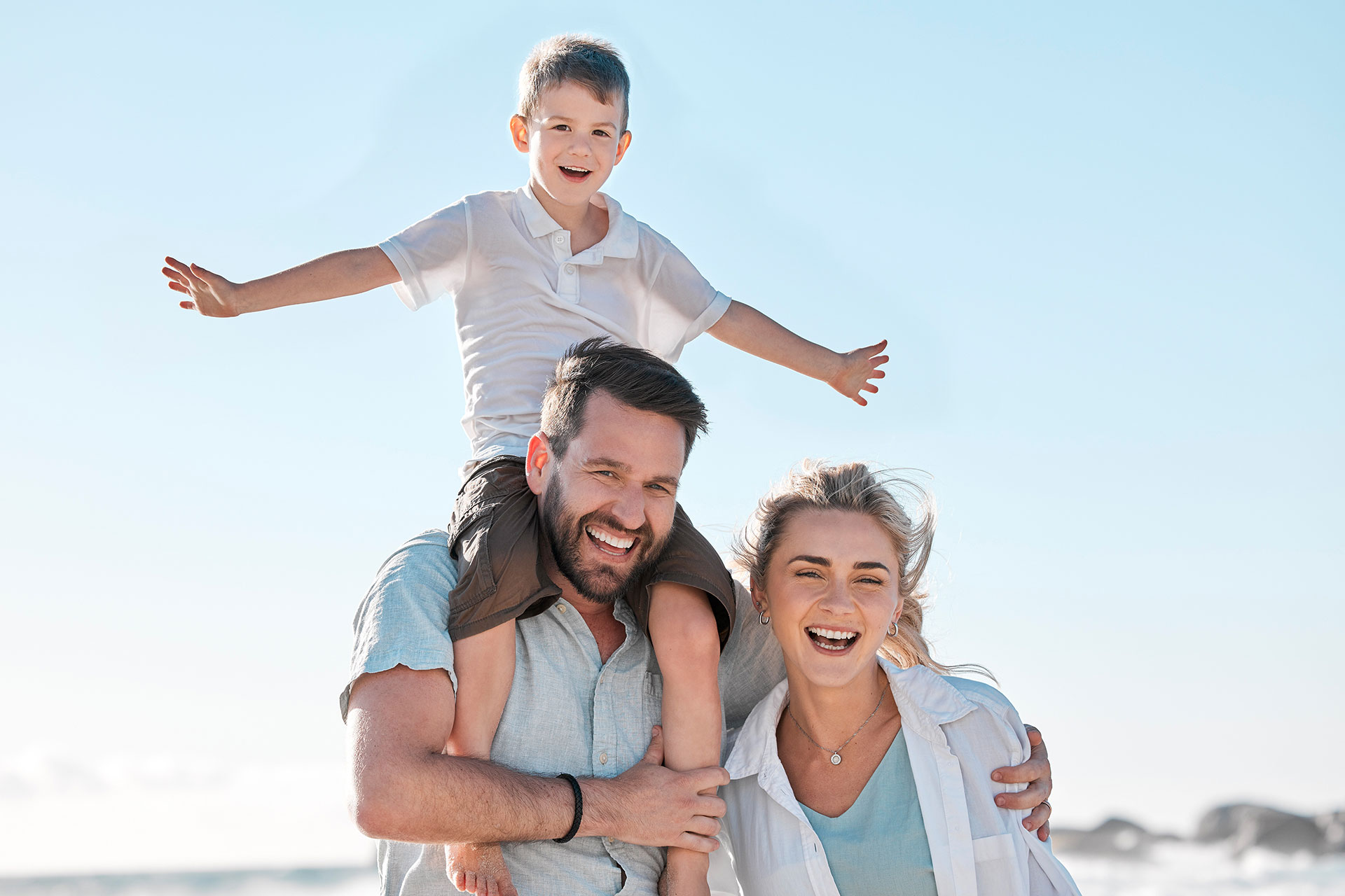 Família tranquila na praia, com pai carregando o filho nas costas e esposa ao lado, representando segurança e bem-estar